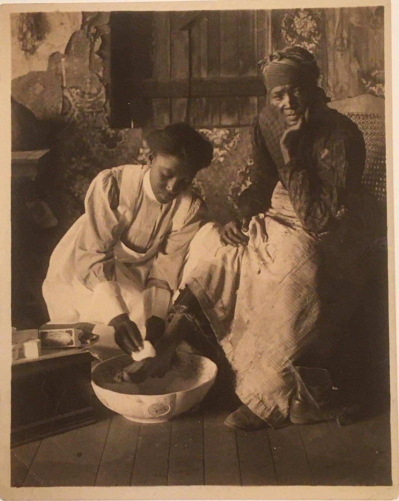 Black and White photograph of a young African American nurse washing an older woman's foot, circa 1920.